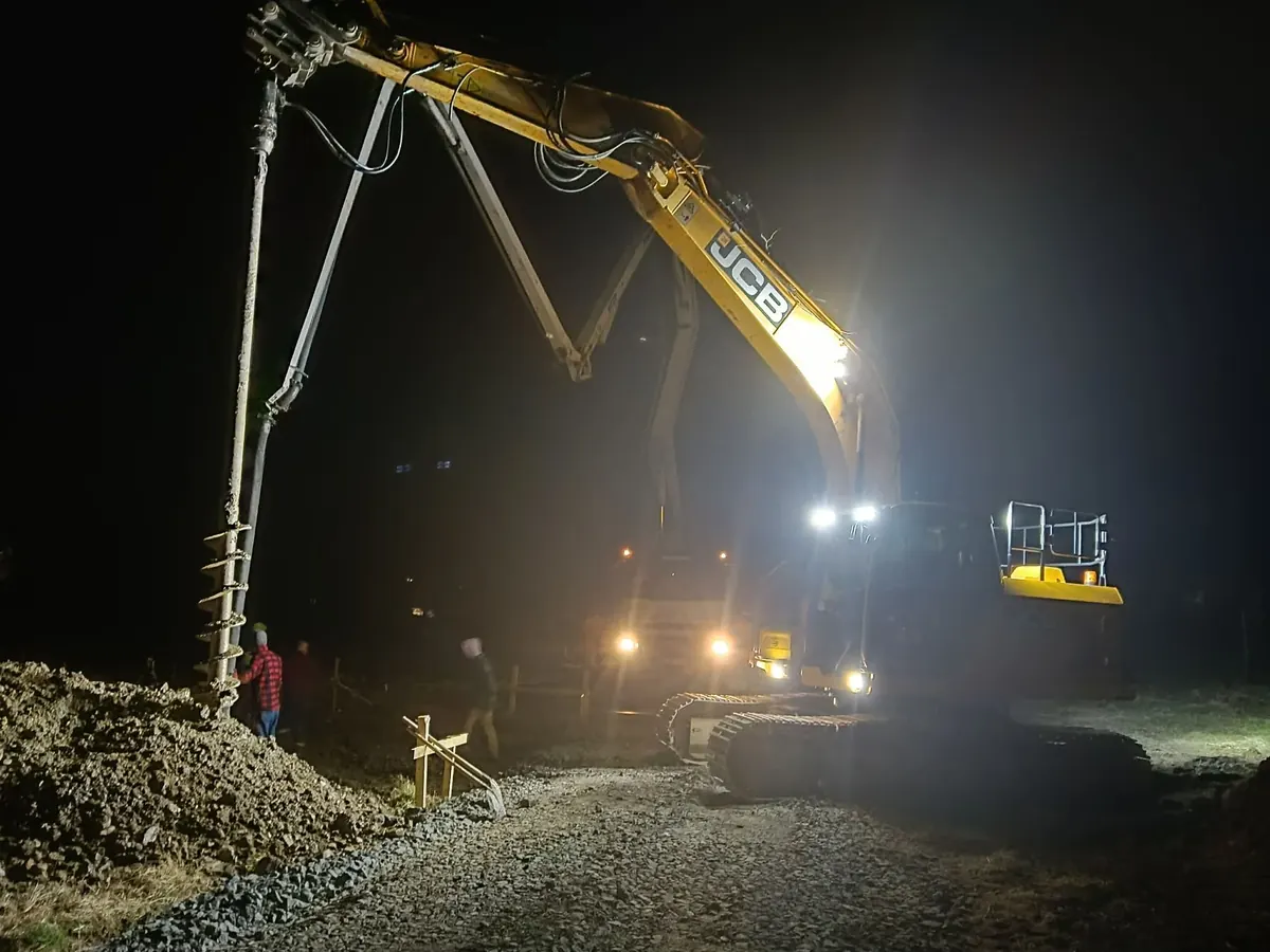 Koparka JCB z osprzętem wiertniczym podczas wiercenia otworów pod pale przy oświetleniu roboczym